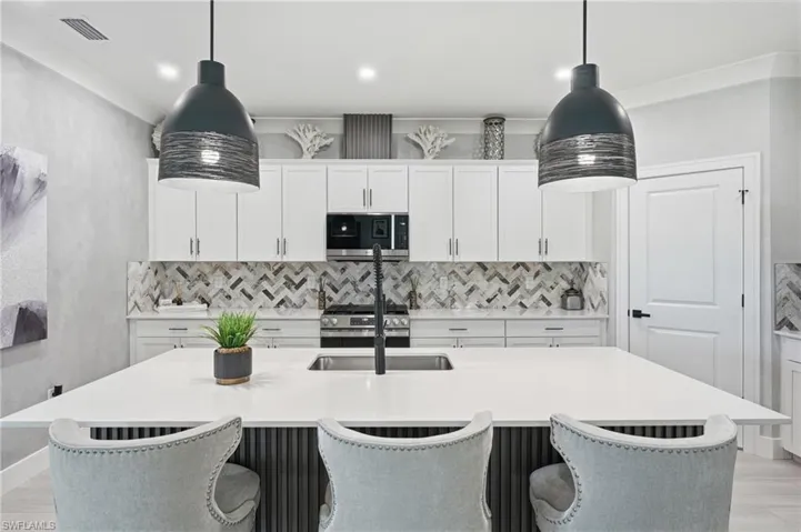 Contemporary kitchen featuring white cabinetry with brushed nickel hardware, a geometric marble tile backsplash, stainless steel appliances including a built-in range, quartz waterfall island countertop with seating for three, and pendant lighting with metal shades suspended above the workspace