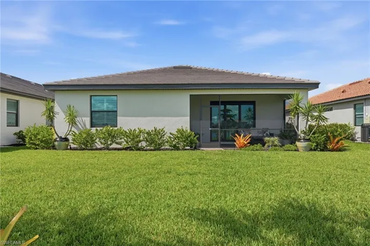 Contemporary home with clean architectural lines and a modern aesthetic, featuring a wide front-facing porch with glass sliding doors, large windows with dark frames, and a manicured lawn bordered by tropical landscaping including palms and flowering plants