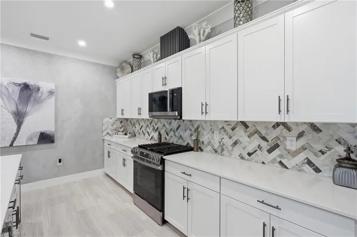 Sleek galley kitchen featuring white cabinetry with brushed metal hardware, quartz countertops, a geometric marble tile backsplash, stainless steel gas range, and built-in microwave