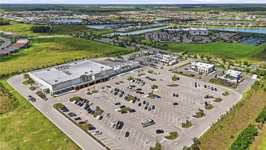 Modern commercial facility featuring an expansive asphalt parking lot with organized striping and landscaped perimeter plantings, a large warehouse structure with a light-colored roof and contemporary architecture, and additional administrative buildings positioned across the manicured grounds