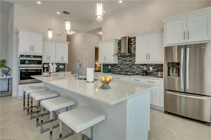 Kitchen featuring appliances with stainless steel finishes, white cabinetry, an island with sink, wall chimney range hood, and pendant lighting