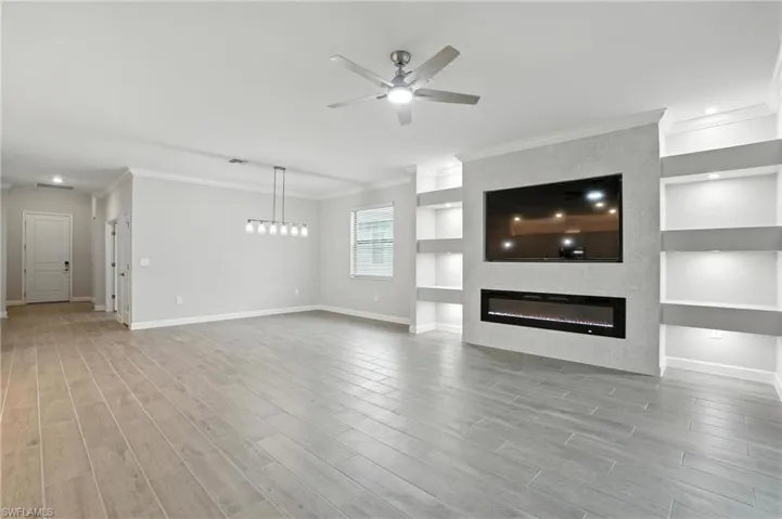 Unfurnished living room with a ceiling fan, built in features, a tiled fireplace, wood finished floors, and crown molding