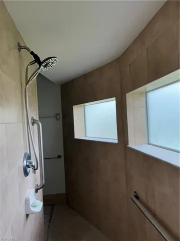 Spa-inspired primary bathroom featuring dual showerheads with handheld fixture, frameless glass enclosure, linear grab bars, and expansive tile surfaces in neutral earth tones