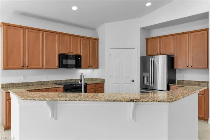 Kitchen featuring light tile patterned floors, a kitchen bar, brown cabinets, black appliances, and light stone counters