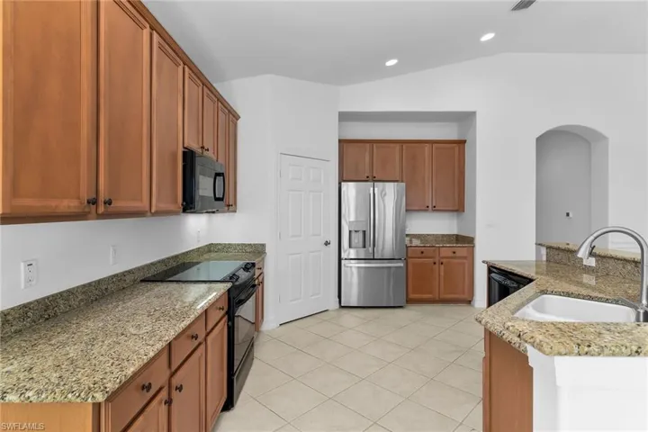 Kitchen with black appliances, light stone counters, brown cabinetry, light tile patterned floors, and arched walkways.