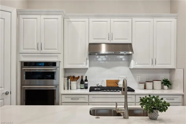 Kitchen with decorative backsplash, range hood, stainless steel appliances, light stone countertops, and white cabinetry