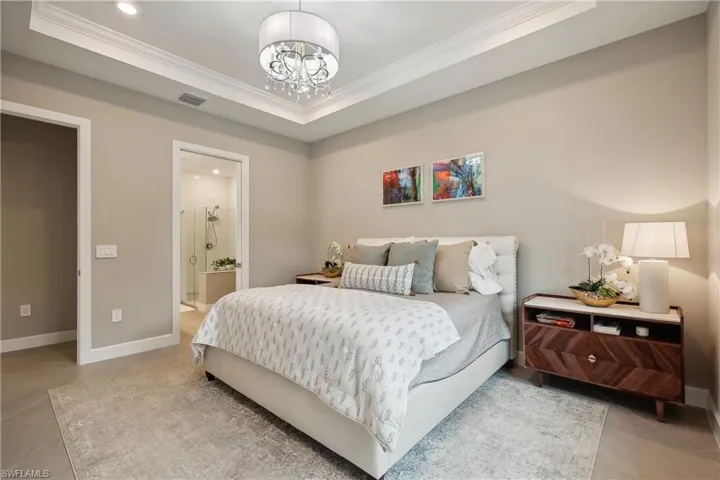 Bedroom with light tile patterned flooring, a raised ceiling, a notable chandelier, ornamental molding, and connected bathroom
