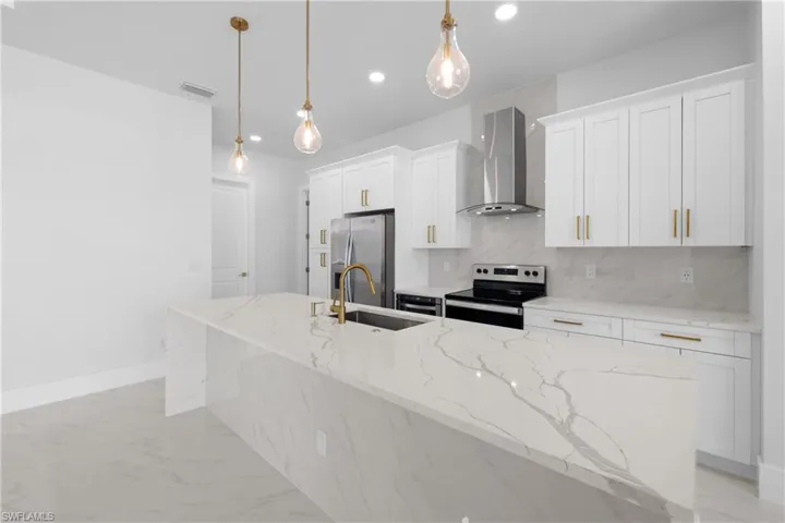 Kitchen with appliances with stainless steel finishes, wall chimney exhaust hood, light stone countertops, and an island with sink