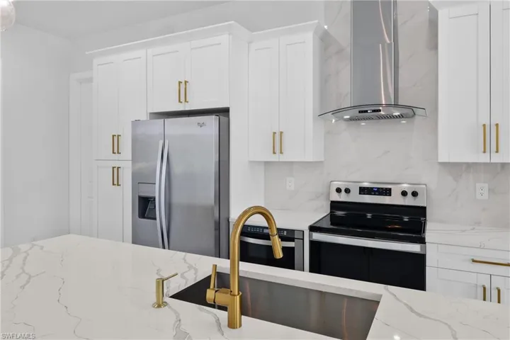 Kitchen with light stone countertops, appliances with stainless steel finishes, wall chimney range hood, and white cabinetry