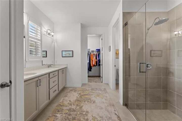 Luxurious primary bath with linen closet, PLANTATION SHUTTERS, double sink vanity with drawer stack,QUARTZ COUNTER with under mount sinks, STAGGERED TILE FLOOR, large shower with tile surround and FRAMELESS SHOWER GLASS DOOR, and water closet.