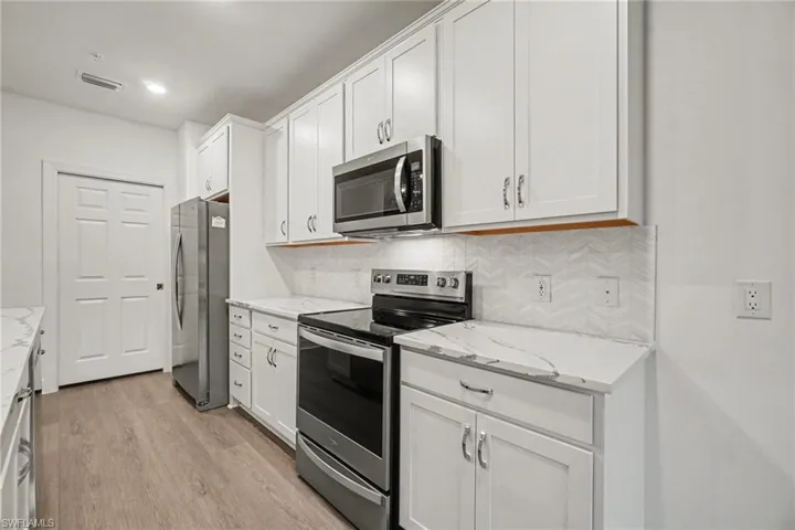 Kitchen featuring white shaker-style cabinetry, stainless steel appliances, marble-look countertops, a herringbone pattern tile backsplash, and wood-finish flooring