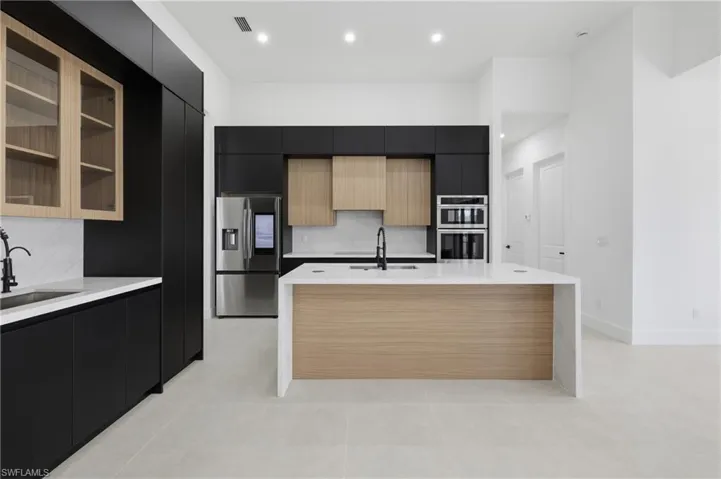 Kitchen featuring dark cabinetry, modern cabinets, glass insert cabinets, appliances with stainless steel finishes, and a center island with sink
