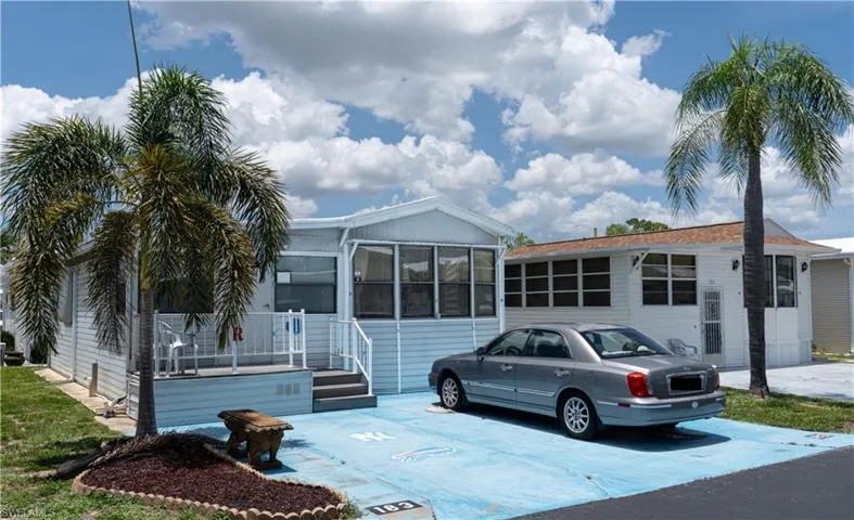 White mobile home featuring a covered porch with white railings, a front palm tree, a blue-painted concrete driveway, and a small landscaped bed