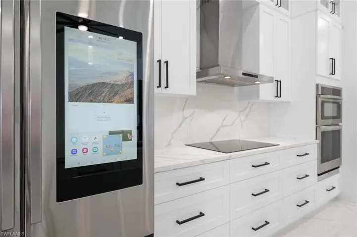 Kitchen with black electric stovetop, white cabinets, light stone counters, wall chimney exhaust hood, and double oven