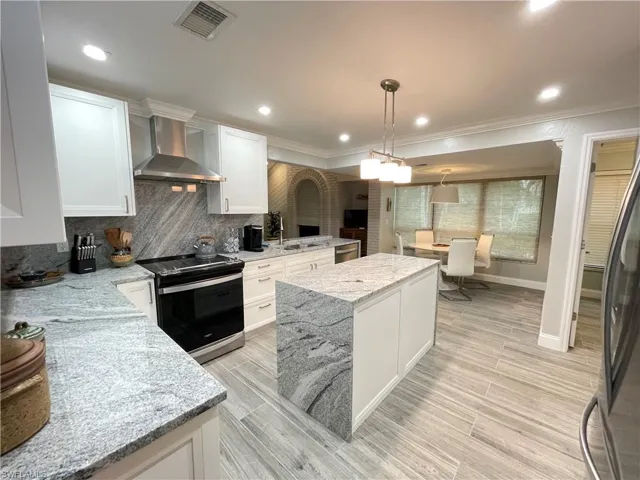 Kitchen featuring hanging light fixtures, white cabinetry, light stone counters, wall chimney range hood, and stainless steel appliances