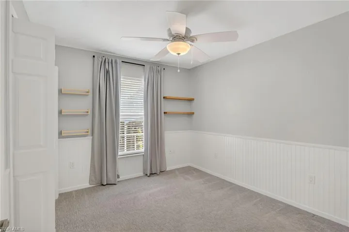 Carpeted interior room featuring a window with blinds and drapes, ceiling fan with light fixture, and wainscoting with wall shelving