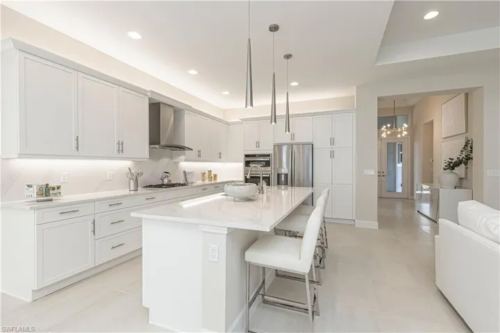 Kitchen featuring hanging light fixtures, wall chimney range hood, a center island with sink, white cabinets, and appliances with stainless steel finishes