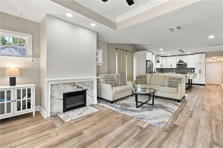 Open-concept living space with wood-finish flooring, a contemporary electrical fireplace featuring a marble surround, and a kitchen with white cabinetry and stainless steel appliances