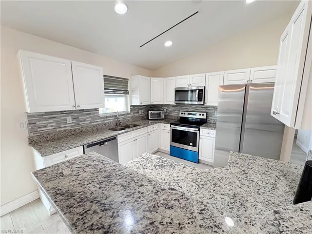 Kitchen featuring stainless steel appliances, lofted ceiling, light stone counters, white cabinets, and backsplash