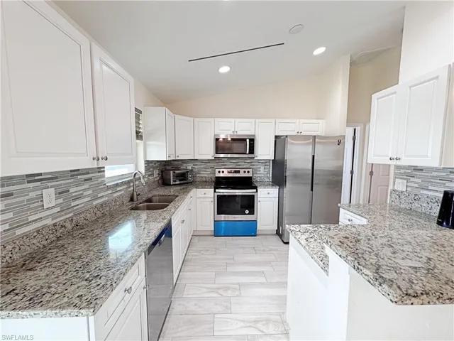 Kitchen with tasteful backsplash, stainless steel appliances, vaulted ceiling, white cabinetry, and light stone counters
