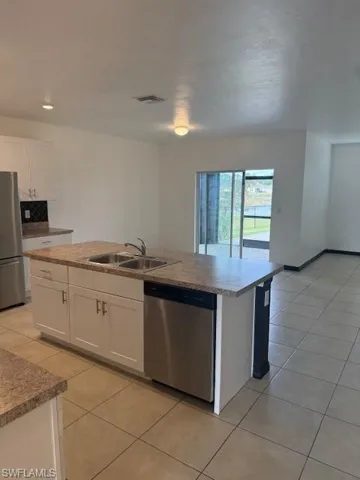 Kitchen with white cabinetry, a center island with sink, stainless steel dishwasher, refrigerator, and light tile patterned floors