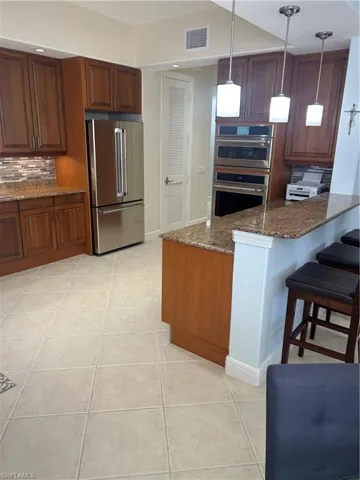 Kitchen featuring dark stone countertops, stainless steel fridge, light tile patterned floors, a kitchen bar, and white double oven