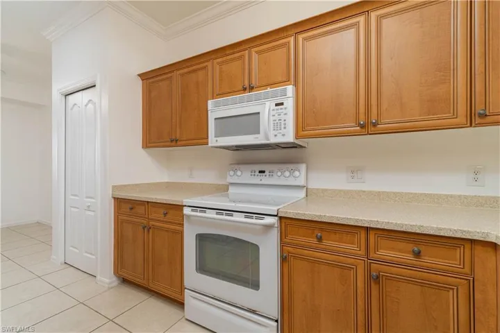 Kitchen featuring light countertops, white appliances, light tile patterned flooring, crown molding, and brown cabinetry