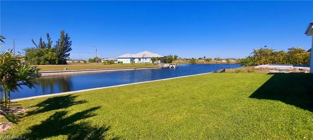 The property features a well-maintained lawn, palm trees, and a clear view of the water under a bright blue sky