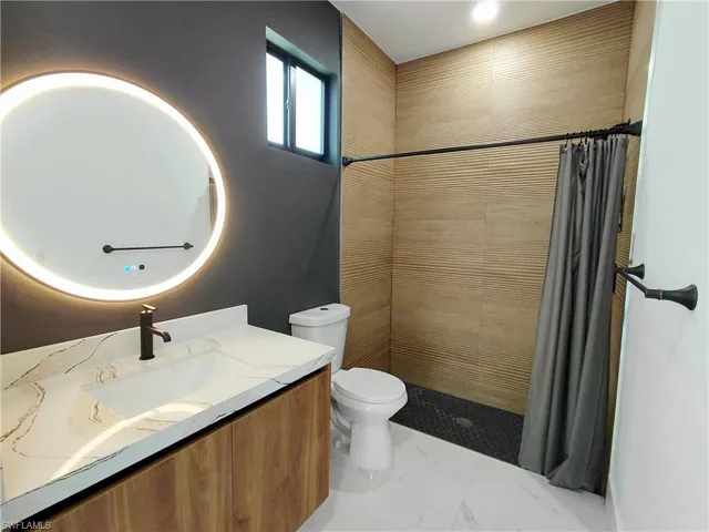 The bathroom features a modern vanity with a white countertop and rectangular sink, a circular backlit mirror, and a shower area with textured tan wall tiles and a dark tiled floor
