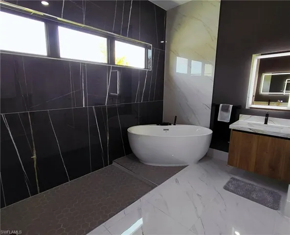 Luxurious bathroom featuring a freestanding soaking tub, large format black and white marbled wall tiles, a floating wood vanity with a marbled countertop, and an illuminated vanity mirror