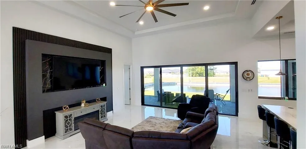 The living area features a modern accent wall with an integrated entertainment space, high ceilings with recessed lighting and a ceiling fan, and large sliding glass doors leading to an outdoor area with a water view