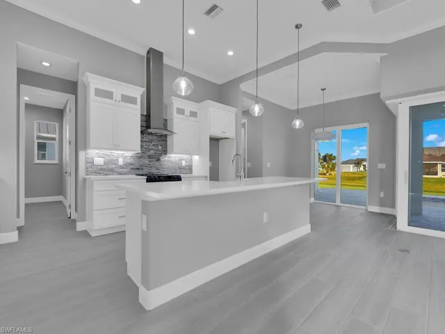 Kitchen with wall chimney range hood, a large island with sink, light countertops, crown molding, and decorative backsplash