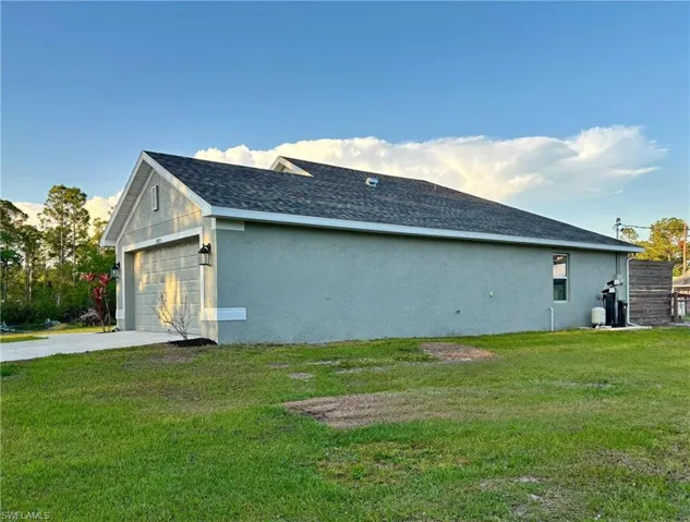 Back of house with roof with shingles, concrete driveway, stucco siding, and a yard