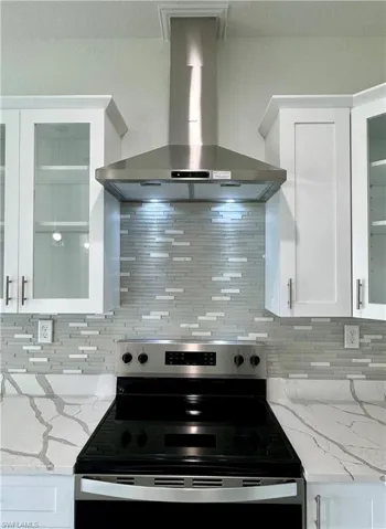 Kitchen with wall chimney range hood, light stone countertops, glass insert cabinets, and white cabinetry
