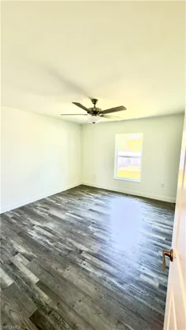 Spacious room featuring wood-finish flooring, a ceiling fan with integrated lighting, a single window, white baseboards, and a white interior door with bronze hardware