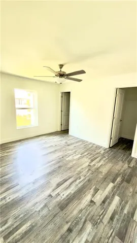 Spacious room featuring wood-finish flooring, a ceiling fan with light fixture, white walls, and a window for natural light