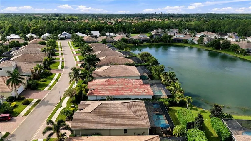 Aerial perspective of a residential community featuring a large retention pond, mature landscaping, paved roadways, and homes with tile roofing