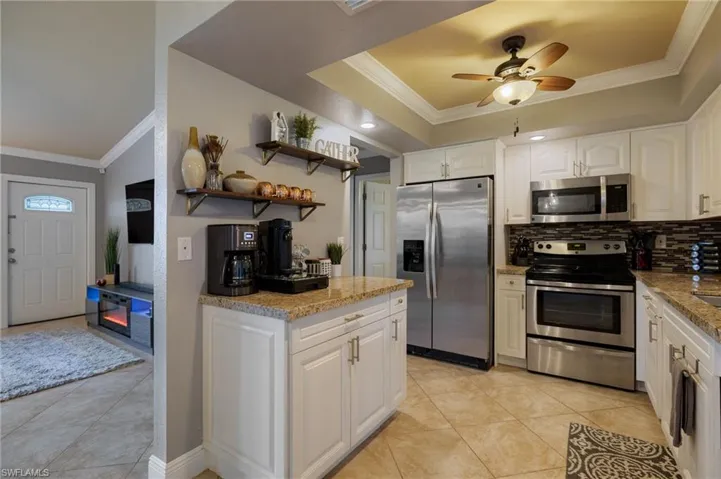 Kitchen with ornamental molding, appliances with stainless steel finishes, white cabinetry, open shelves, and light stone counters