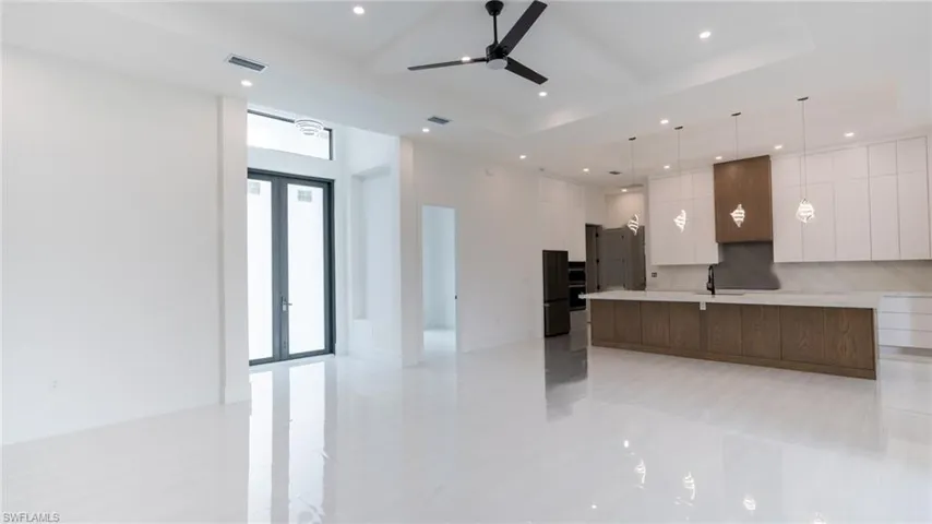 Spacious open-concept living area featuring reflective white tile flooring, a modern kitchen island with pendant lighting, black ceiling fan, recessed lighting, and double-door entry