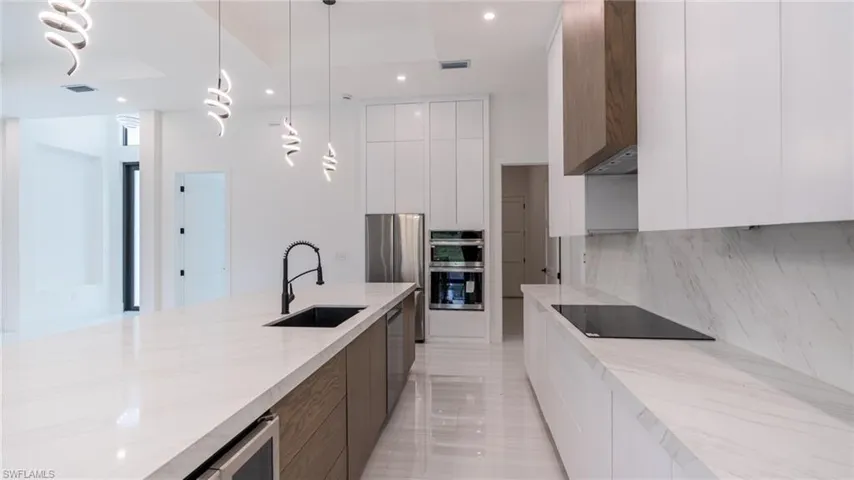 Spacious kitchen featuring a large central island with an undermount sink, quartz countertops, integrated stainless steel appliances, and sleek white cabinetry with wood-finish accents