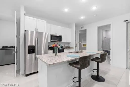 Kitchen featuring appliances with stainless steel finishes, sink, white cabinetry, light stone counters, and a kitchen island with sink