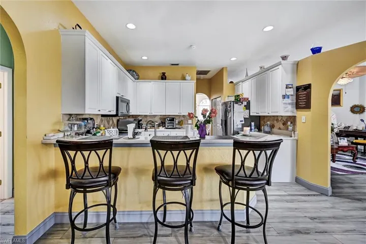 Kitchen with arched walkways, a peninsula, white cabinets, wood-style tile flooring, and recessed lighting. Breakfast bar, tasteful backsplash, white cabinets, and stainless steel fridge.