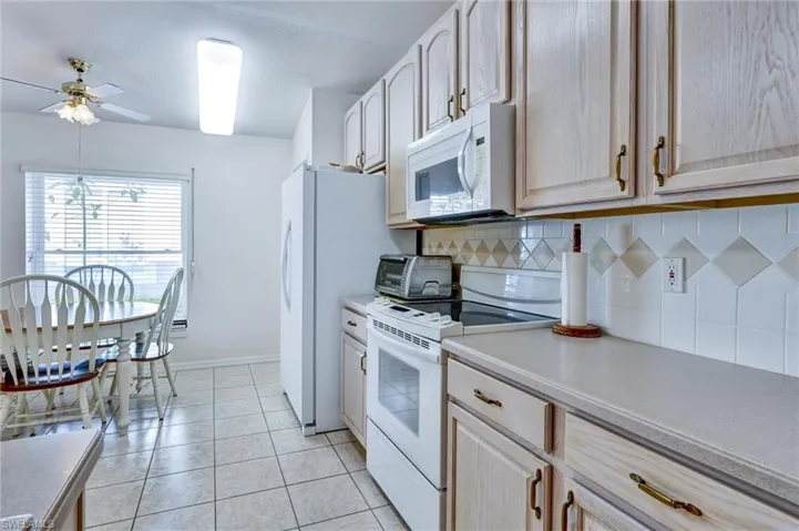 Kitchen featuring light wood-finish cabinetry with brass-tone hardware, white appliances, tiled backsplash with decorative accents, and light-toned countertops