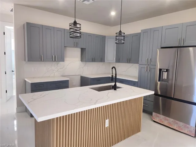 Kitchen featuring stainless steel refrigerator with ice dispenser, light stone countertops, decorative light fixtures, a kitchen island with sink, and backsplash
