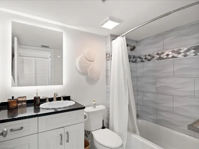 Bathroom featuring a white vanity with dark countertop, white shell-shaped sink, illuminated mirror, and a tiled shower with decorative accent band