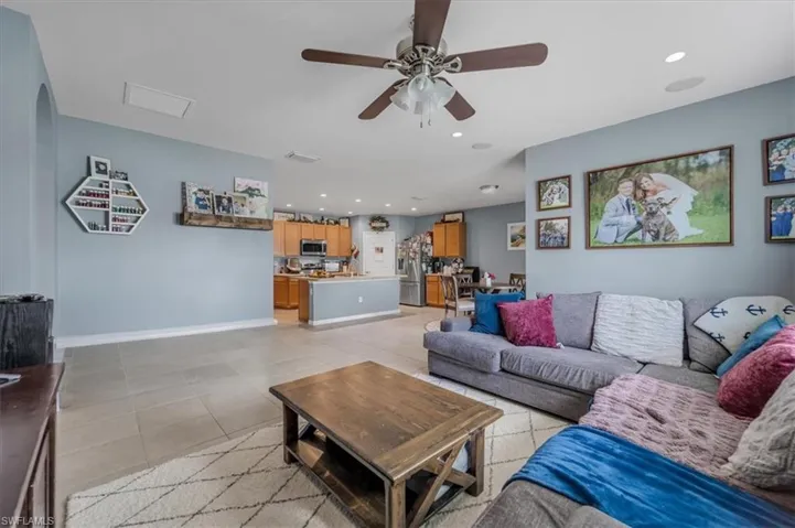 Open-concept living area featuring light neutral tile flooring, a ceiling fan with light fixture, recessed lighting, and a partial view of the kitchen with wood cabinetry and a breakfast bar
