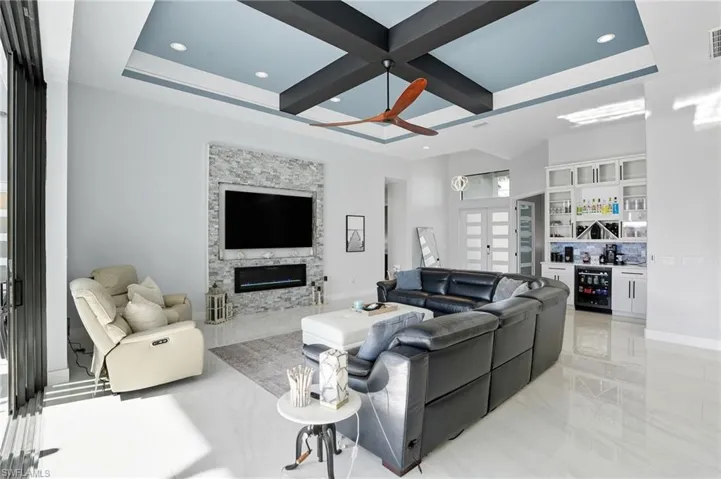 Living room featuring coffered ceiling, a dry bar, a stone fireplace, light marble finish floors, and wine cooler