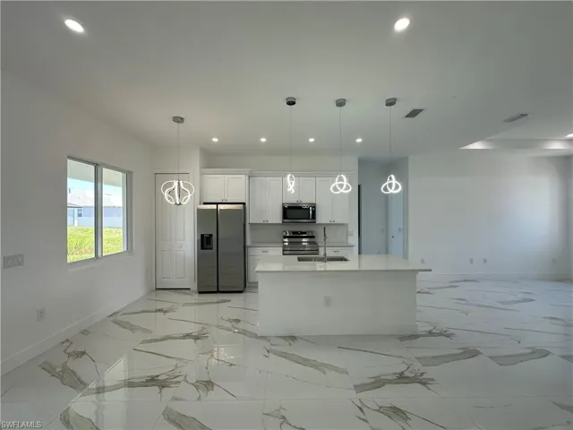 Kitchen with white cabinetry, hanging light fixtures, appliances with stainless steel finishes, light marble finish floors, and an island with sink