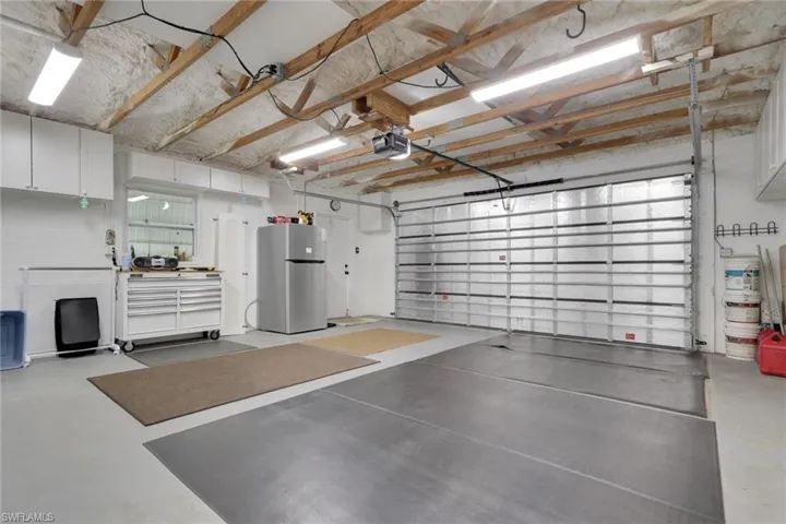 Spacious garage featuring exposed wood beam ceiling, multiple fluorescent light fixtures, upper wall cabinetry, and a large roll-up garage door