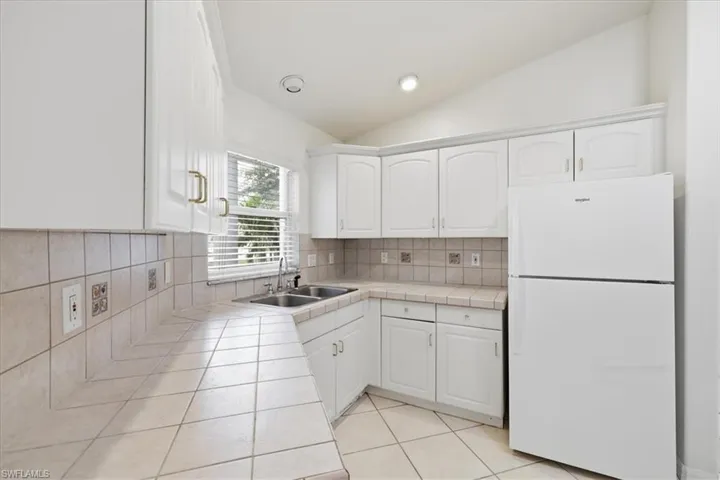 Kitchen featuring white cabinetry with gold-toned hardware, a tile backsplash, tile countertops, and a double basin sink beneath a window with blinds